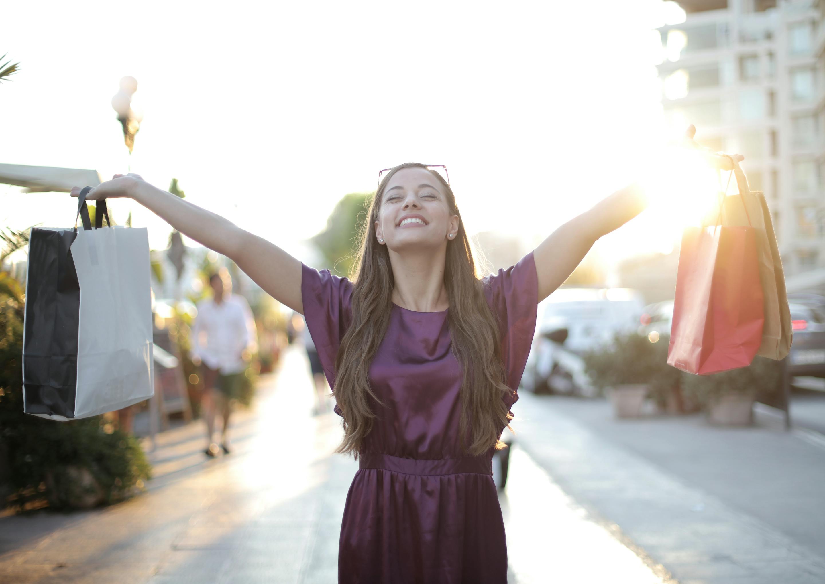 Cheerful woman with shopping bags enjoying sunny day, arms raised in excitement.