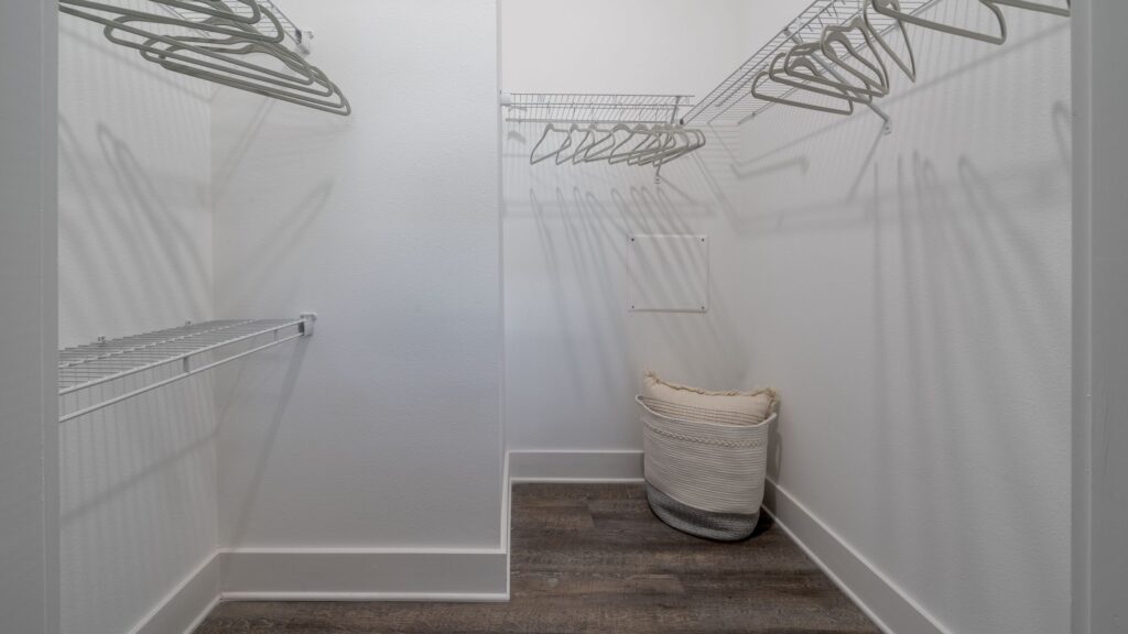 Empty walk-in closet with white walls, wire shelves, hangers, and a laundry basket on the wooden floor.