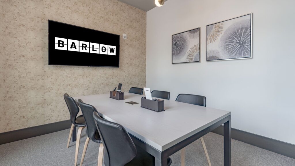 A modern meeting room with a rectangular table, six chairs, two wall art pieces, and a screen displaying the word "BARLOW.