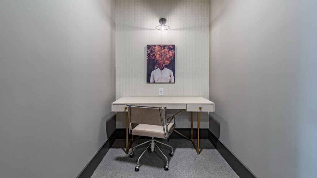 Minimalist workspace with a desk and chair under a wall lamp, featuring a framed art piece of a person with flowers instead of a head.