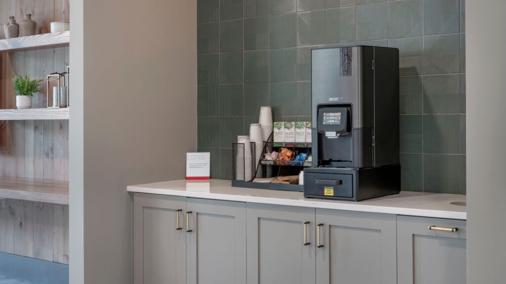 Coffee machine on a countertop with cups, lids, and coffee supplies against a tiled wall.