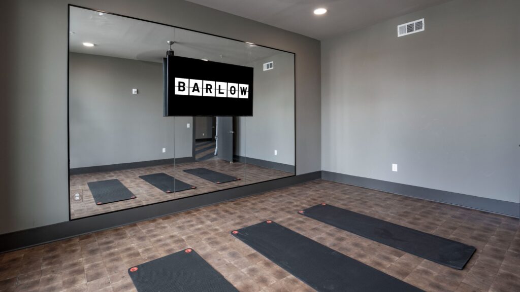 Room with exercise mats on a tiled floor, a large mirror on the wall, and a screen displaying the word "BARLOW.