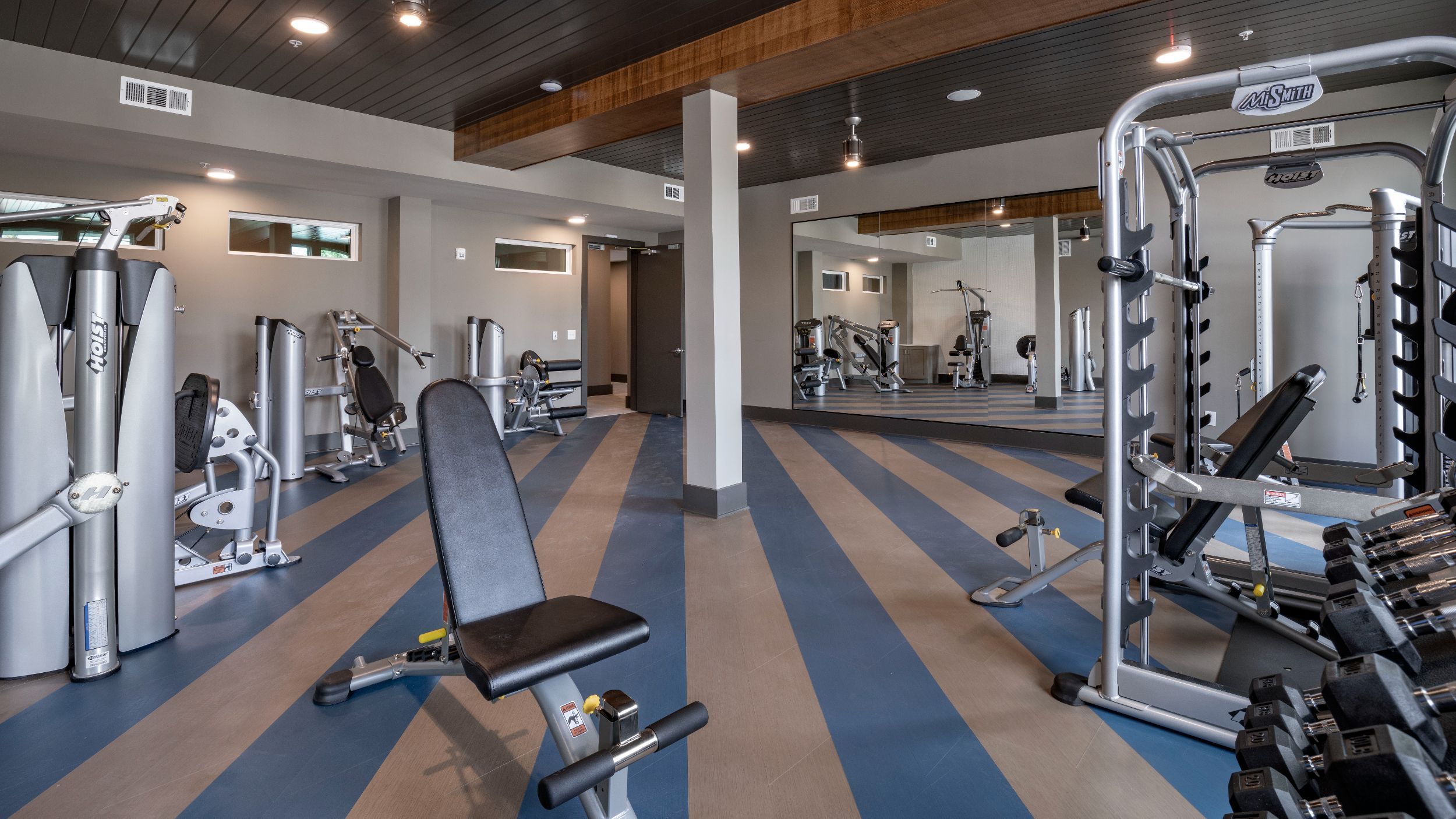 Modern gym with a variety of exercise equipment, including weight machines, benches, and free weights. A large mirror covers one wall, and the floor has blue and gray stripes.