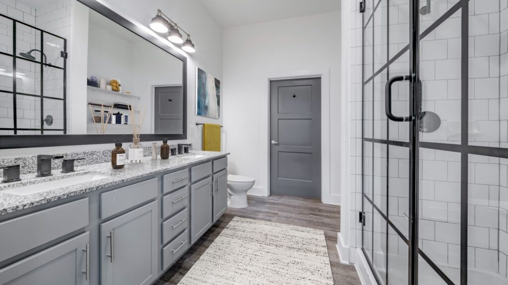 Modern bathroom with a double sink vanity, granite countertop, glass shower enclosure, and a gray door. Wall-mounted lights and decorative items are visible, with wood flooring and a rug.