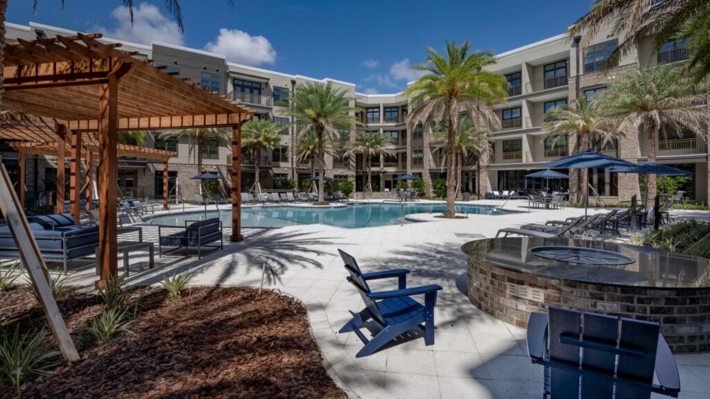 Outdoor pool area with lounge chairs, umbrellas, and palm trees surrounded by a modern multi-story building. A wooden pergola and a round fire pit with chairs are in the foreground.