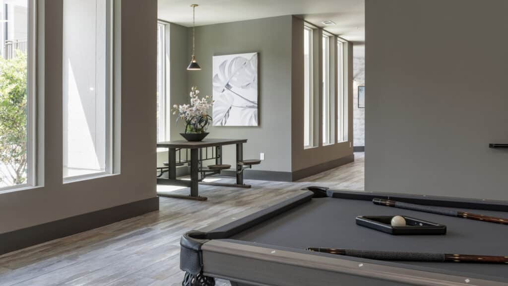 A modern room with a black pool table in the foreground and a dining table with benches and a potted plant near large windows in the background.