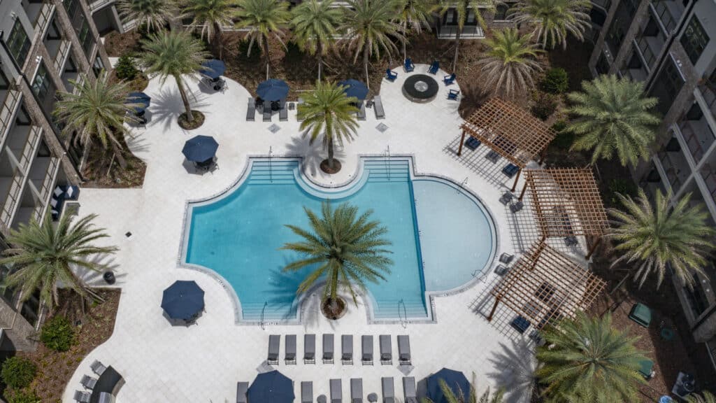 Aerial view of a rectangular outdoor swimming pool surrounded by lounge chairs, umbrellas, palm trees, pergolas, and landscaped areas in an apartment or hotel courtyard.